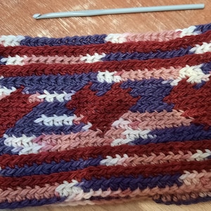 May include: A handmade crochet cowl with a heart pattern. The cowl uses burgundy, white, pink, and blue yarn. A silver crochet hook is placed on the wooden surface beside the cowl. The cowl is approximately 25 cm wide.