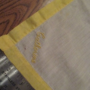 May include: A white handkerchief with a yellow border and the name "Catherine" embroidered in yellow thread.