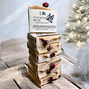 May include: A stack of six handmade soap bars with a label that reads "Patchouli Blush Patchouli Essential Oil Handmade Soap Enriched with Organic Shea & Cocoa Butter, Raw Coconut Oil with French Pink Clay & Natural Yogurt Handmade in Small Batches 90 g". The soap bars are a light beige colour and have dried rose buds on top.