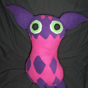 Puede incluir: Un juguete de peluche con forma de criatura con orejas moradas, cuerpo rosa con patrones de diamantes morados y ojos verdes.