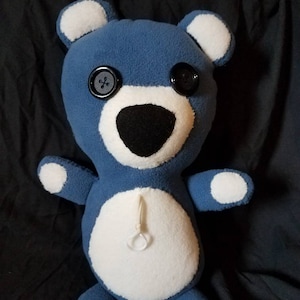 以下が含まれることがあります： 白いアクセントと黒いボタンの目をつけた青いぬいぐるみクマ。クマは白いお腹に白い紐が白い輪に繋がっています。