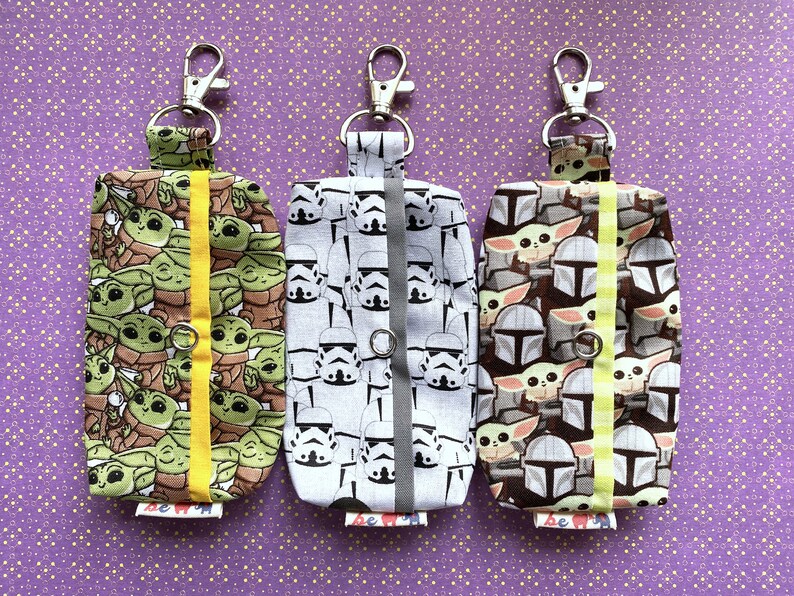 Puede incluir: Tres llaveros de tela con diferentes dise&ntilde;os de personajes de Star Wars. El llavero de la izquierda presenta un patr&oacute;n repetido de Baby Yoda, el llavero del medio presenta un patr&oacute;n repetido de Stormtroopers y el llavero de la derecha presenta un patr&oacute;n repetido de personajes mandalorianos.