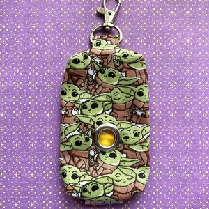 Puede incluir: Un porta bolsas de caca de tela verde y marr&oacute;n con un clip plateado y un ojal amarillo. El porta bolsas est&aacute; cubierto con un patr&oacute;n de peque&ntilde;os personajes verdes con ojos grandes.