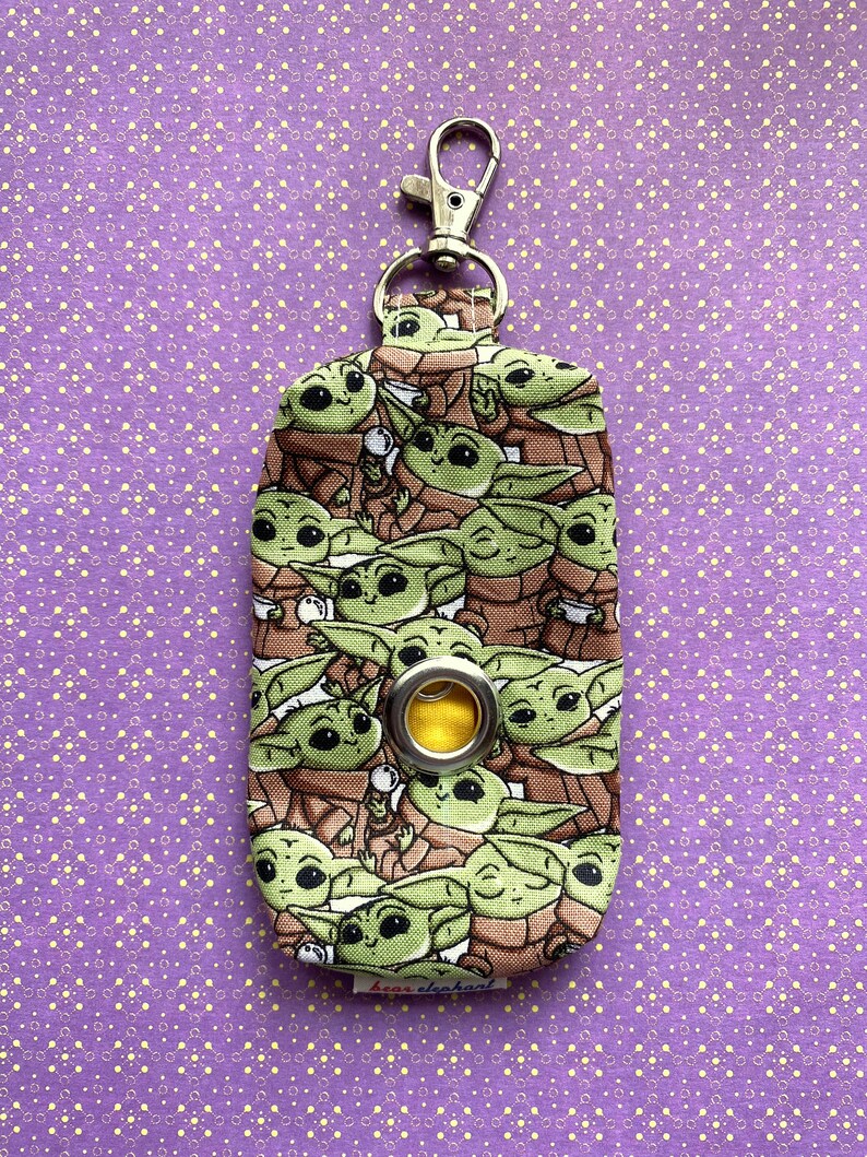 Puede incluir: Un porta bolsas de caca de tela verde y marr&oacute;n con un clip plateado y un ojal amarillo. El porta bolsas est&aacute; cubierto con un patr&oacute;n de peque&ntilde;os personajes verdes con ojos grandes.