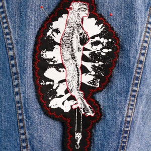 May include: A denim jacket with a large, embroidered patch. The patch shows a black and white illustration of a figure with a red outline, framed by a black border with red stitching. Two pins are placed on the jacket.