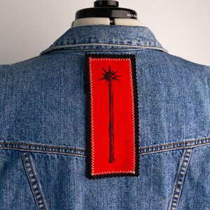May include: A blue denim jacket featuring a rectangular patch sewn onto the back. The patch is bright red with a black border and a black graphic illustration of a spiked mace centred on the fabric, attached with white zigzag stitching.