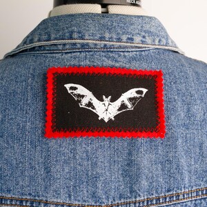 May include: A light blue denim jacket featuring a rectangular black patch on the back. The patch displays a white graphic of a bat with spread wings and is bordered by a vibrant red fabric edge with a decorative scalloped stitch pattern.
