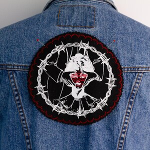 May include: A blue denim jacket with a circular patch on the back. The patch has a black background with a white and red graphic of a person with barbed wire and a heart-shaped design. Two pins are visible.