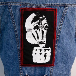 May include: A light blue denim jacket with a large patch on the back. The patch has a white gas mask graphic on a black background, with a red border. The jacket is a classic style.