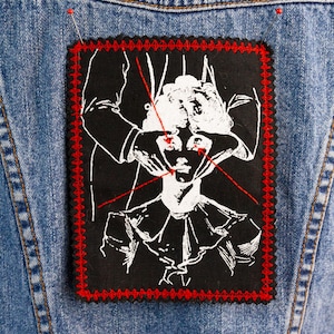 May include: A black patch with a white illustration of a person's face being manipulated by hands. The patch has a red stitched border and is pinned to a denim jacket. The illustration features red lines and a ruffled collar.