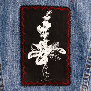 May include: A black rectangular patch with a white floral design and red scalloped edges is pinned to a denim jacket. The floral design features a large flower and delicate stems.