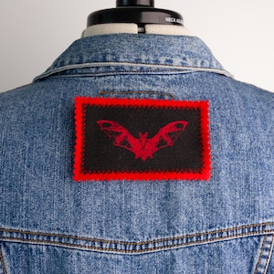 May include: A light blue denim jacket featuring a rectangular black patch on the back. The patch displays a red bat graphic and is bordered by a vibrant red zigzag stitched edge, contrasting against the textured blue fabric of the denim garment.