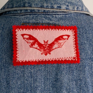 May include: A blue denim jacket featuring a rectangular patch sewn onto the back. The patch has a light pink background with a red bat graphic, framed by a red border with a zigzag stitched edge, centred below the jacket collar.