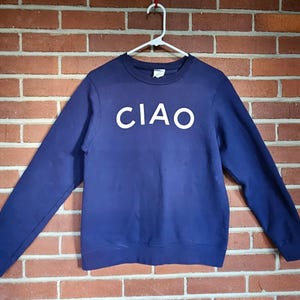 May include: A navy blue sweatshirt with the word "CIAO" in white block letters across the chest. The long-sleeved top is hanging on a white hanger against a brick wall.