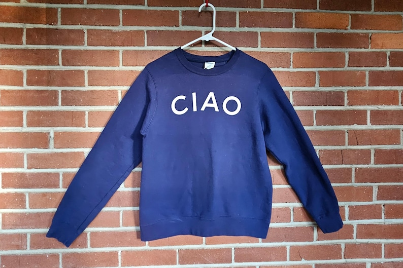 May include: A navy blue sweatshirt with the word "CIAO" in white block letters across the chest. The long-sleeved top is hanging on a white hanger against a brick wall.
