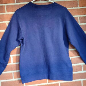 May include: A solid navy blue sweatshirt with long sleeves, displayed against a brick wall. The crew neck sweatshirt is hanging on a white hanger. The back of the sweatshirt is visible, showing the neckline and shoulder area.
