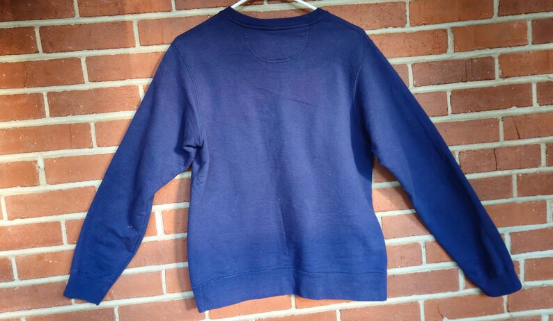 May include: A solid navy blue sweatshirt with long sleeves, displayed against a brick wall. The crew neck sweatshirt is hanging on a white hanger. The back of the sweatshirt is visible, showing the neckline and shoulder area.
