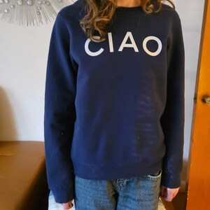 May include: Navy blue sweatshirt with the word "CIAO" in large white letters across the chest. The sweatshirt is a crew neck style and appears to be made of a soft material. The person is wearing blue jeans.