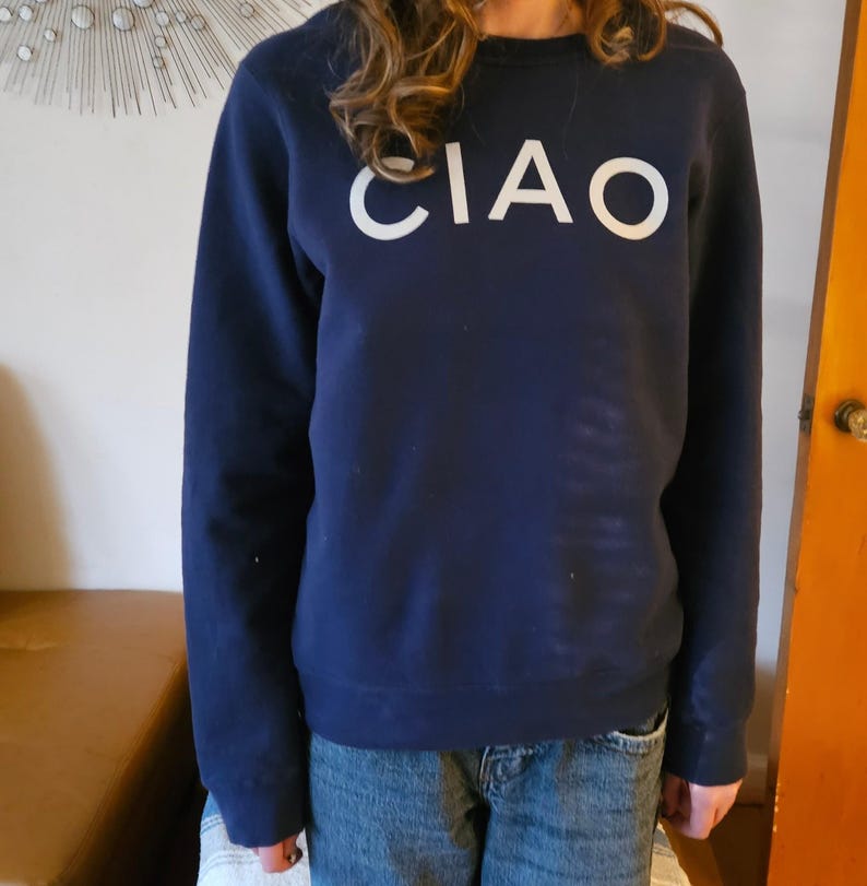 May include: Navy blue sweatshirt with the word "CIAO" in large white letters across the chest. The sweatshirt is a crew neck style and appears to be made of a soft material. The person is wearing blue jeans.