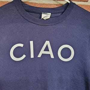 May include: Navy blue crew neck sweatshirt with the word "CIAO" in white block letters across the chest. The sweatshirt is hanging on a white hanger against a brick wall.