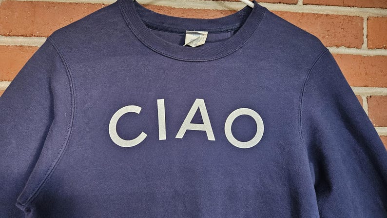 May include: Navy blue crew neck sweatshirt with the word "CIAO" in white block letters across the chest. The sweatshirt is hanging on a white hanger against a brick wall.