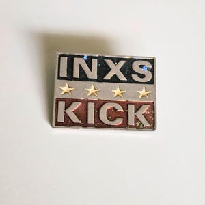 May include: A square pin featuring the text "INXS" above four gold stars and the word "KICK" in a red and silver design. The pin has a silver border and is set against a white background.