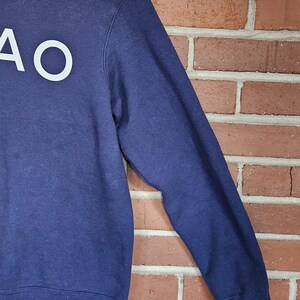 May include: Navy blue sweatshirt with the white letters "AO" on the front. The long-sleeved top is hanging on a white hanger against a red brick wall.
