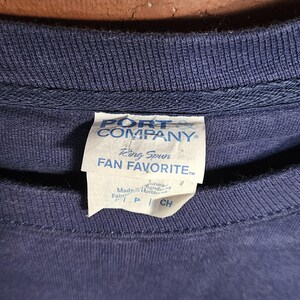 May include: Navy blue crew neck sweatshirt with a tag that reads "PORT COMPANY" and "Ring Spun FAN FAVORITE". The tag also indicates the garment was made in Honduras. The bottom of the shirt has the letters "IA" in white.