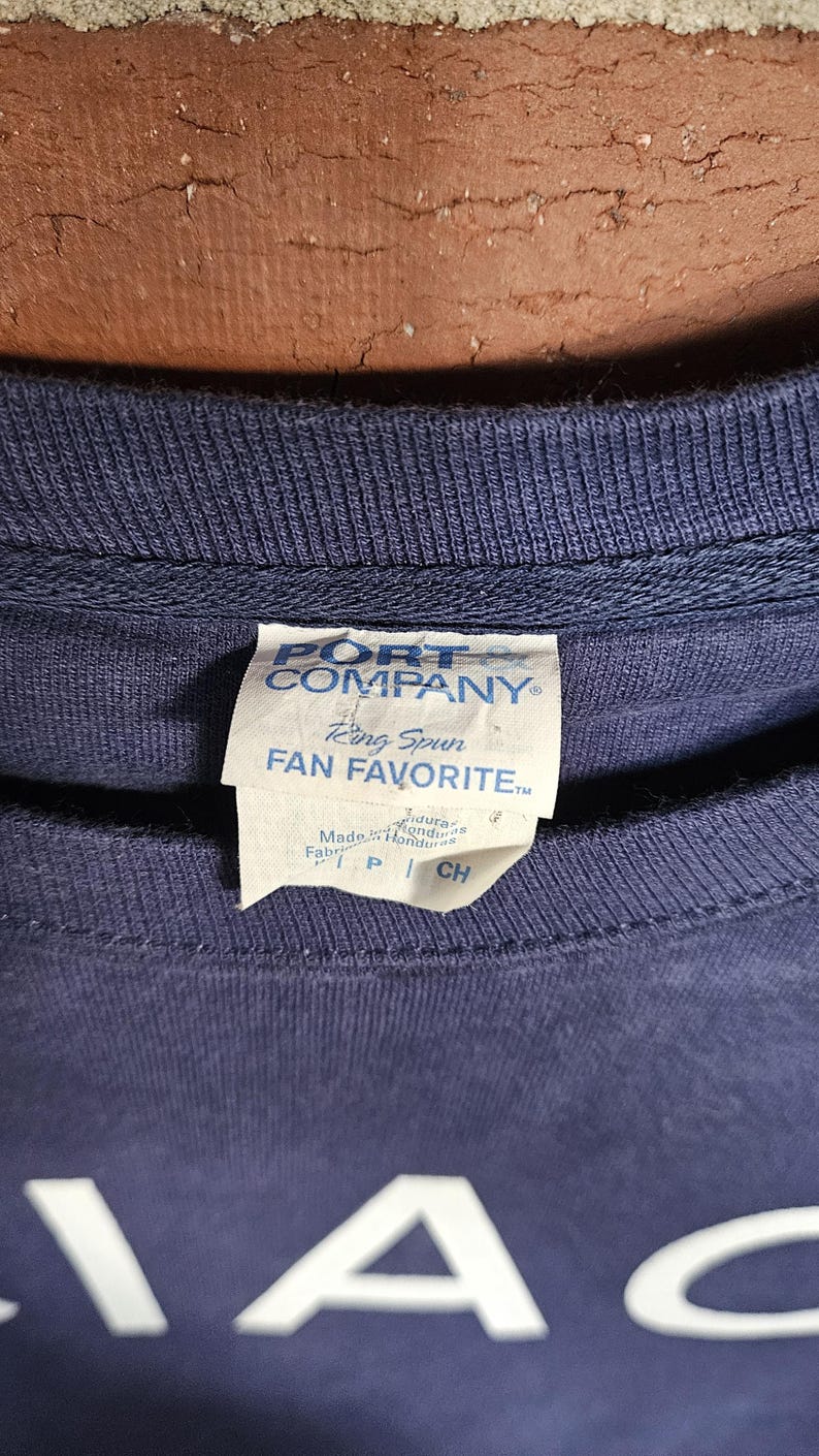 May include: Navy blue crew neck sweatshirt with a tag that reads "PORT COMPANY" and "Ring Spun FAN FAVORITE". The tag also indicates the garment was made in Honduras. The bottom of the shirt has the letters "IA" in white.