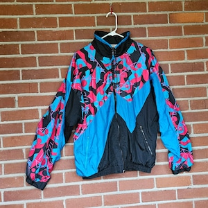 May include: A vintage zip-up jacket with a vibrant geometric pattern in pink, blue, and black. The jacket features a high collar, a full-length zipper, and a black base with blue and pink accents. The jacket is hanging against a brick wall.