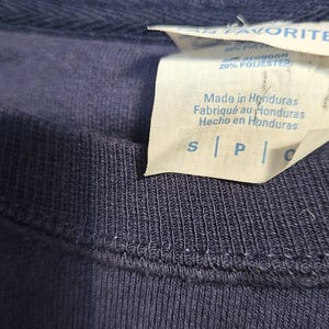 May include: Close-up of a navy blue crew neck shirt. A white tag displays "Made in Honduras" in English, French, and Spanish. The tag also lists the fabric composition as 80% cotton and 20% polyester.