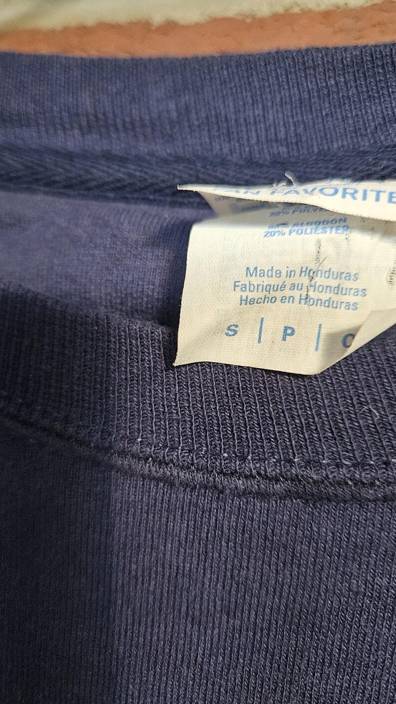 May include: Close-up of a navy blue crew neck shirt. A white tag displays "Made in Honduras" in English, French, and Spanish. The tag also lists the fabric composition as 80% cotton and 20% polyester.