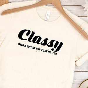 May include: A white t-shirt with black text that reads "Classy with a hint of don't try me tho". The shirt is hanging on a wooden hanger and is surrounded by other items, including a straw hat, sandals, and a cup of coffee.