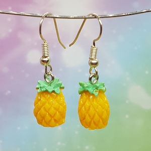 Könnte beinhalten: Ein Paar Ohrringe mit kleinen Ananas-Anhängern. Die Ohrringe haben silberfarbene Haken und gelbe Ananas-Anhänger mit grünen Blättern. Der Hintergrund ist ein weicher, mehrfarbiger Farbverlauf.