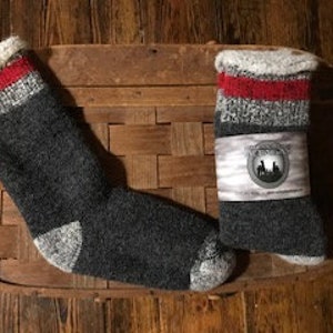 May include: A pair of dark grey wool socks with red and white stripes at the cuff. The socks are displayed on a brown woven basket. One sock has a paper band with a circular emblem.
