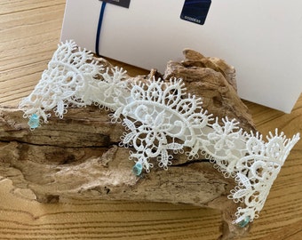 May include: White lace garter with delicate floral patterns and light blue embellishments. The garter is displayed on a piece of driftwood.