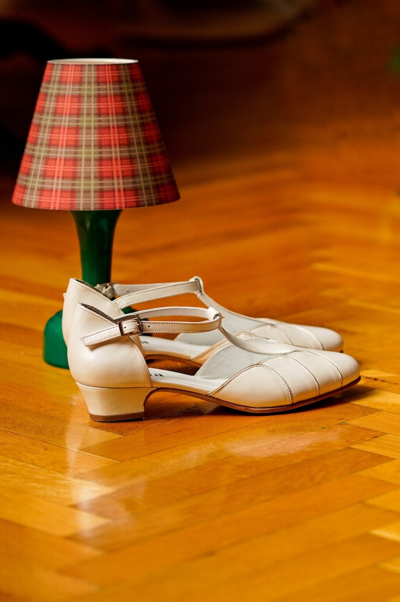 May include: A pair of white leather heeled shoes with ankle straps. The shoes are on a wooden floor with a plaid lampshade in the background.