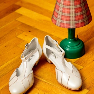 May include: A pair of white leather shoes with a T-strap design. The shoes have a small heel and are labeled "Irma" on the inside of each shoe. The shoes are sitting on a wooden floor next to a green lamp with a red and white plaid lampshade.