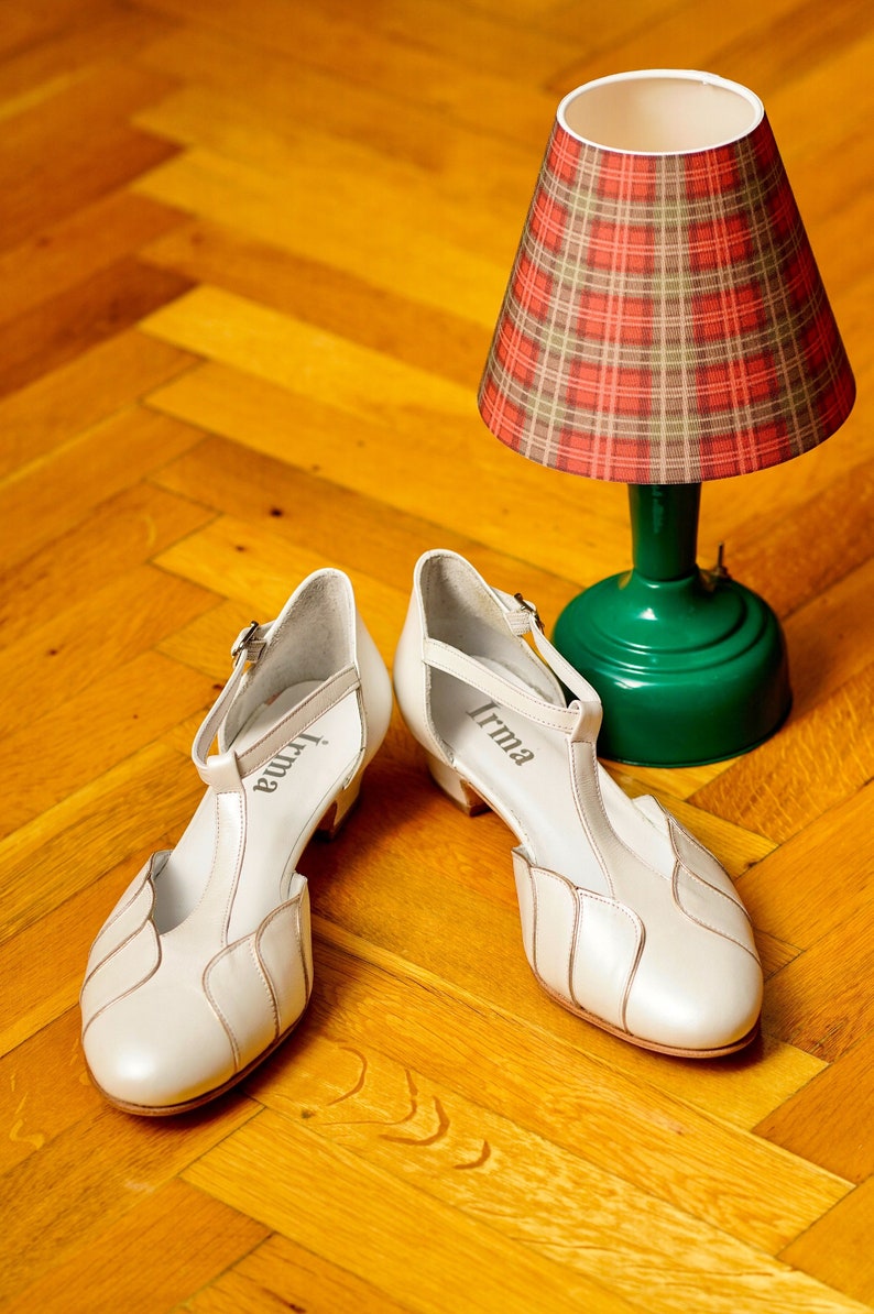 May include: A pair of white leather shoes with a T-strap design. The shoes have a small heel and are labeled "Irma" on the inside of each shoe. The shoes are sitting on a wooden floor next to a green lamp with a red and white plaid lampshade.