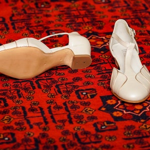 May include: A pair of white leather heeled shoes with ankle straps. The shoes are on a red patterned rug.