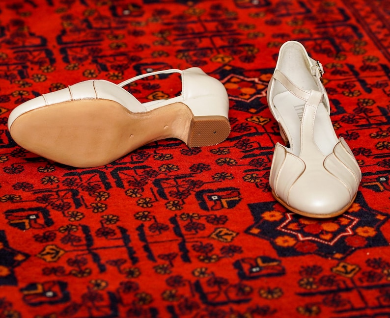 May include: A pair of white leather heeled shoes with ankle straps. The shoes are on a red patterned rug.
