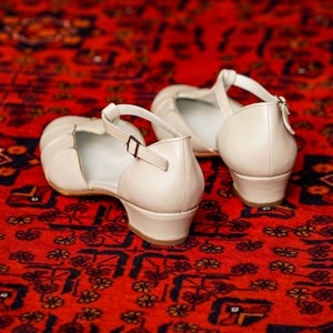 May include: A pair of off-white leather heeled sandals with ankle straps. The sandals are on a red patterned rug.