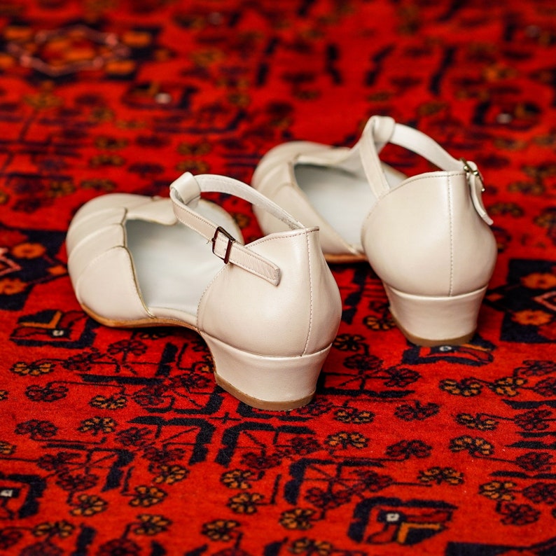 May include: A pair of off-white leather heeled sandals with ankle straps. The sandals are on a red patterned rug.