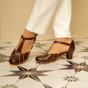 May include: A pair of brown leather heeled shoes with a t-strap design and a scalloped edge. The shoes have a low heel and are being worn by someone with white trousers.
