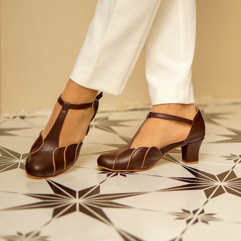May include: A pair of brown leather heeled shoes with a t-strap design and a scalloped edge. The shoes have a low heel and are being worn by someone with white trousers.