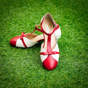 Pu&ograve; includere: Un paio di scarpe da donna rosse e bianche con un fiocco sulla punta e la parola "Irma" all'interno della scarpa.