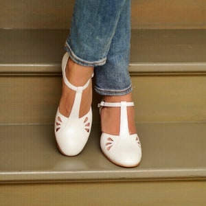 May include: White leather t-strap shoes with cut-out detailing. The shoes are worn by a person wearing blue jeans.