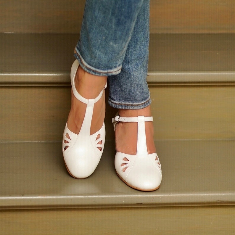 May include: White leather t-strap shoes with cut-out detailing. The shoes are worn by a person wearing blue jeans.