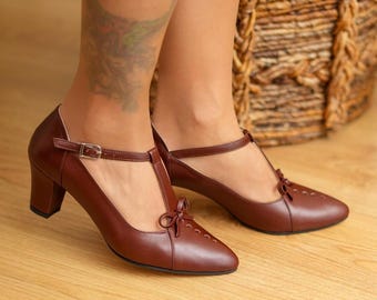 May include: Brown leather T-strap heels with a bow detail on the toe and a buckle closure. The heels have a block heel design. The shoes are a classic style, suitable for various occasions.