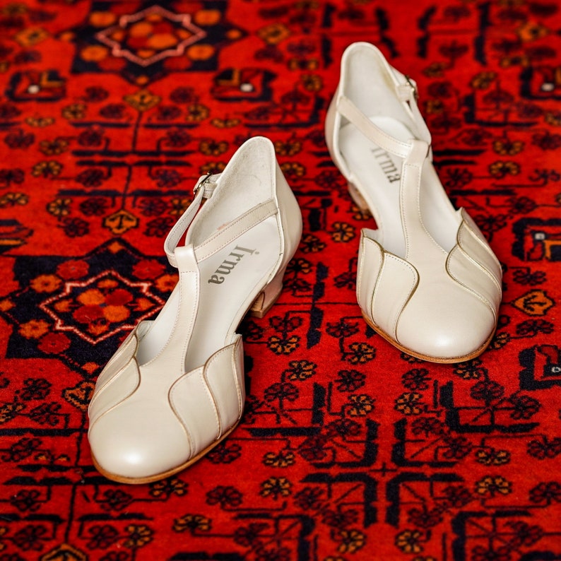 May include: A pair of white leather heeled shoes with a t-strap design. The shoes have a small heel and are decorated with a simple, elegant design. The brand name "Irma" is printed on the inside of each shoe.
